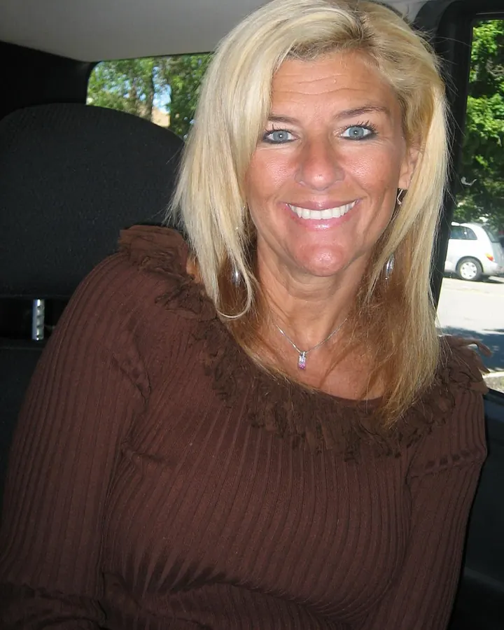 Femme blonde en haut violet, souriante, assise sur un lit.