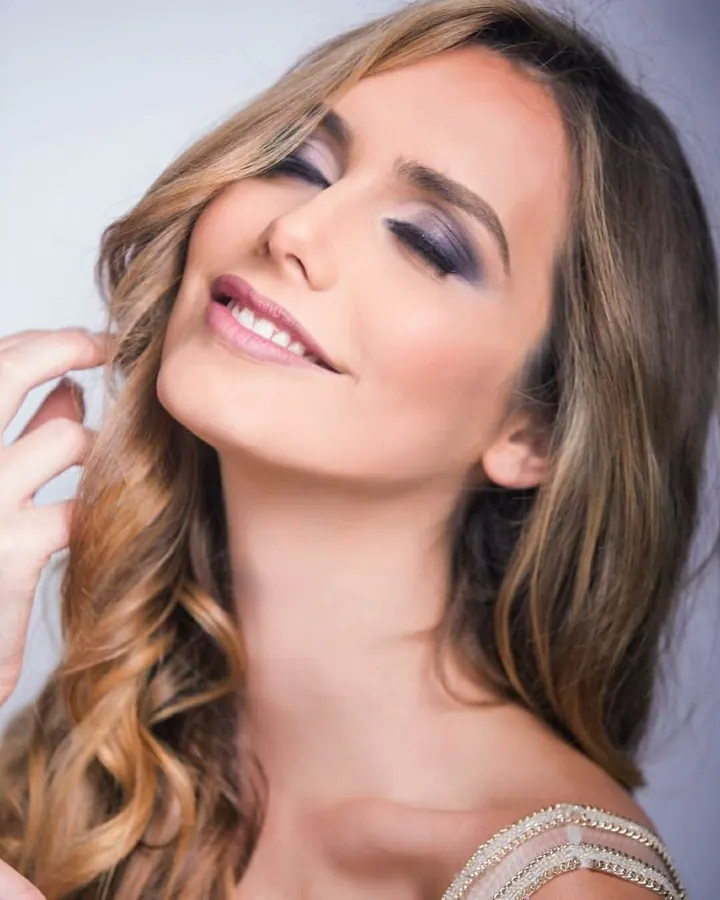 Femme aux cheveux blonds, en haut gris, posant de face, dans une pièce avec bibliothèque