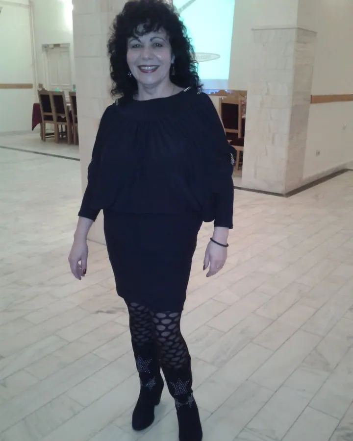 Femme aux cheveux bruns foncés, en tenue noire, debout dans une salle avec sol carrelé, souriante
