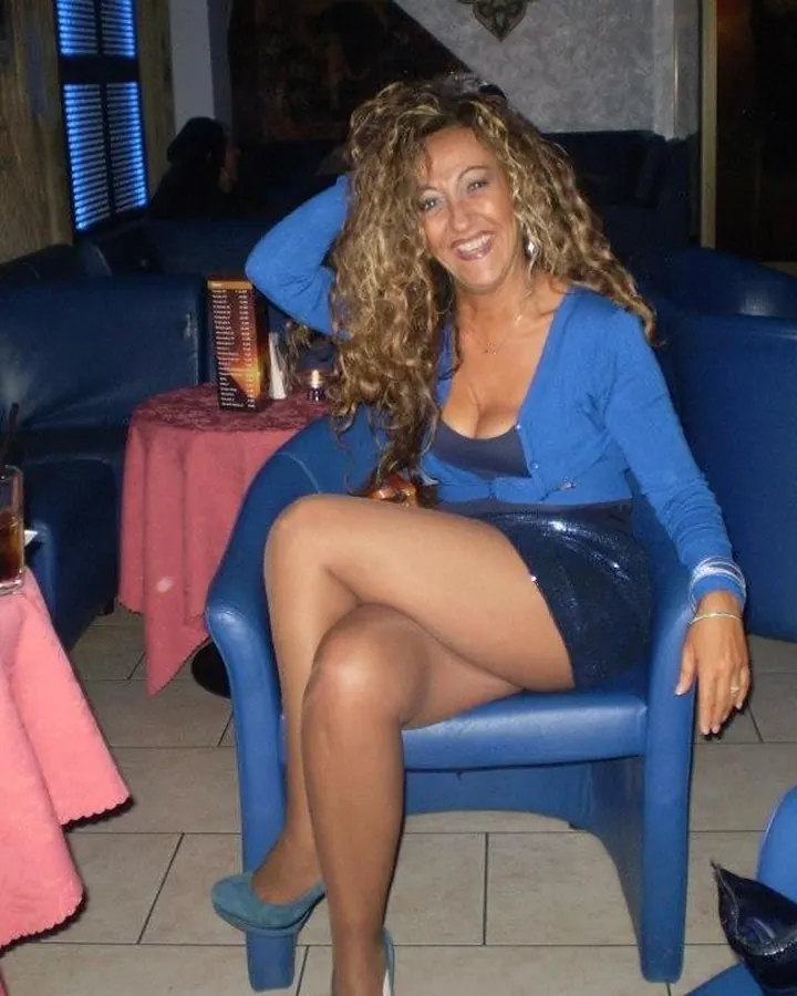 Femme aux cheveux châtains bouclés, assise sur un fauteuil bleu dans un bar, en mini-jupe noire et cardigan bleu, souriante