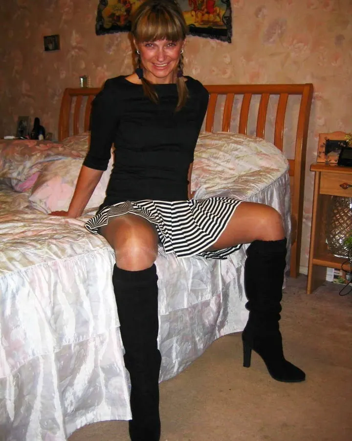 Femme européenne aux cheveux châtains, portant un haut noir et une jupe rayée, assise sur un lit dans une chambre décorée de rose.