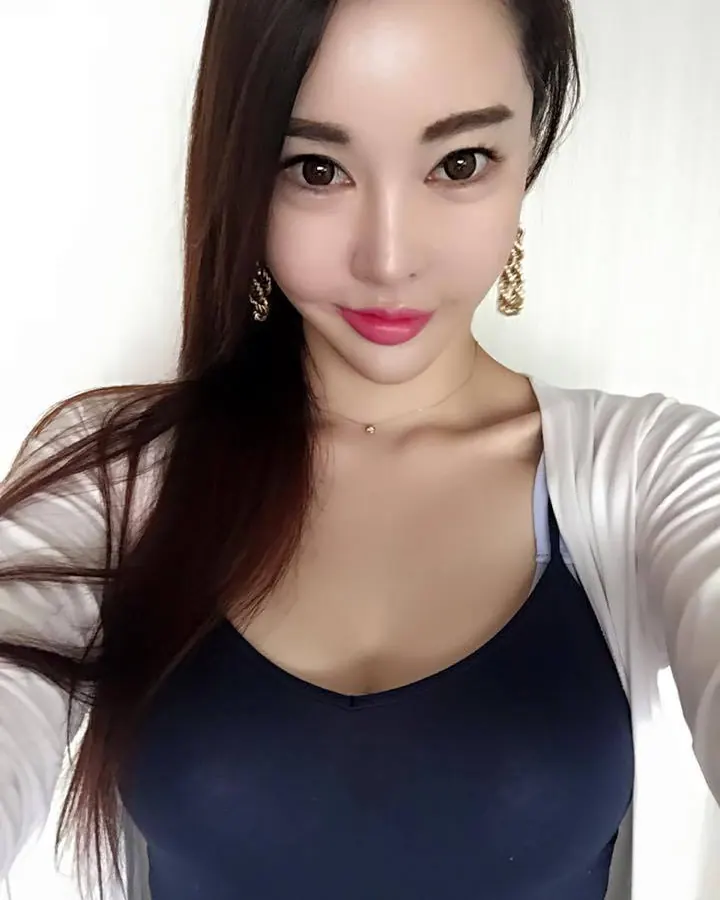 Jeune femme asiatique aux cheveux châtains, portant un haut noir et un short blanc, assise sur un lit avec un sourire.