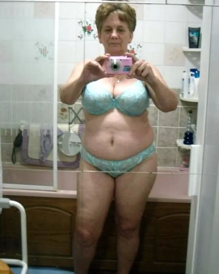 Femme plus âgée aux cheveux blonds, portant un soutien-gorge et un string bleu clair, prenant un selfie dans une salle de bain.