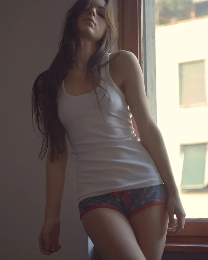 Jeune femme aux cheveux bruns portant un débardeur blanc et des sous-vêtements gris, appuyée contre un mur près d'une fenêtre.