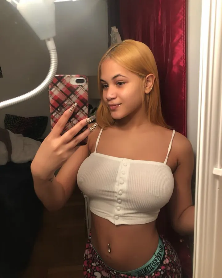 Jeune femme blonde, portant un haut blanc et un short rose, prenant un selfie dans un miroir, cheveux longs, lunettes.