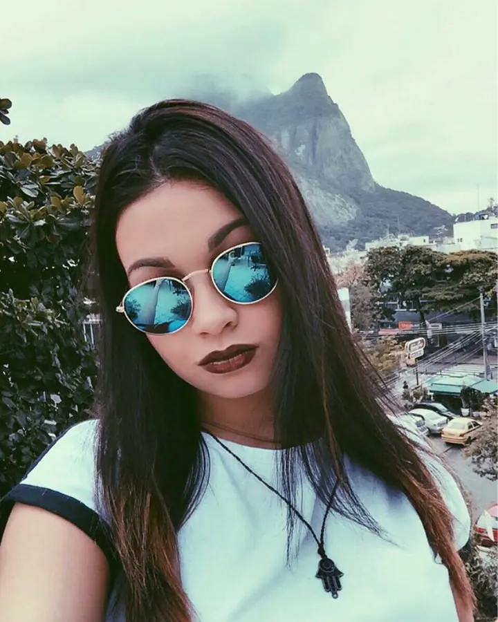 Femme aux cheveux bruns foncés, lunettes de soleil rondes, en t-shirt blanc, prenant un selfie devant une montagne rocheuse.