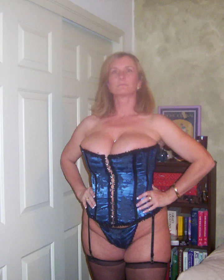 Femme aux cheveux blonds, en corset bleu brillant, collants noirs, debout dans une chambre, les mains sur les hanches.