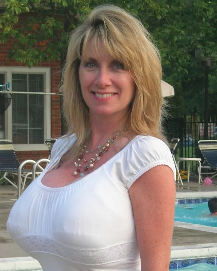 Femme blonde aux yeux bleus, souriante, en haut blanc, debout près d'une piscine avec des chaises longues en arrière-plan.