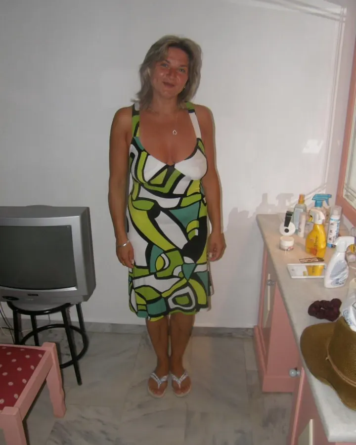 Femme aux cheveux gris, en robe à motifs géométriques verts, blancs et noirs, debout dans une pièce avec un vieux téléviseur et une table de toilette.