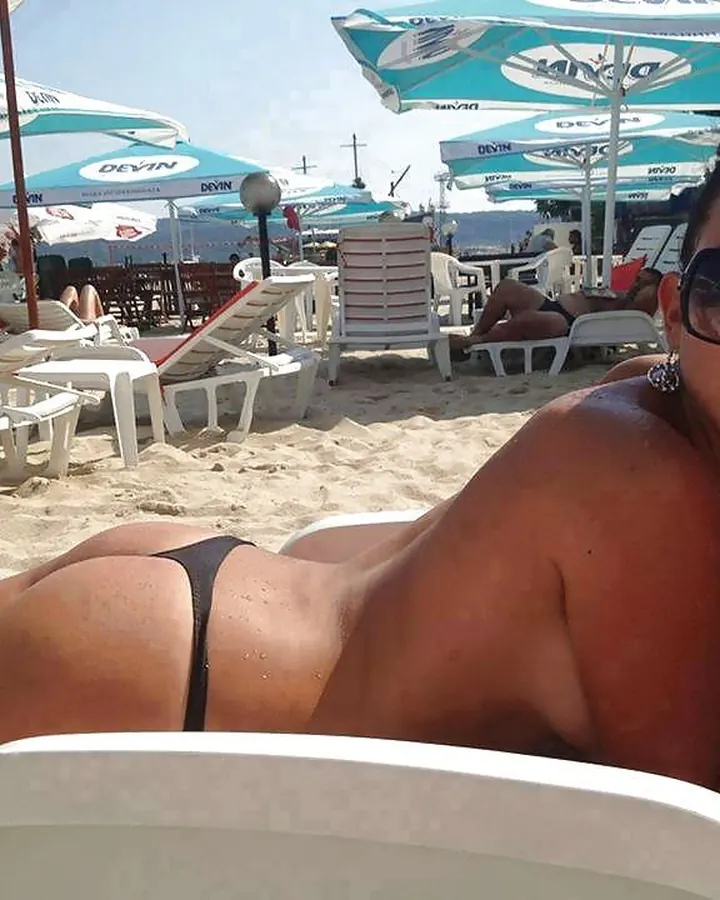 Femme aux cheveux bruns noirs, en maillot de bain noir, allongée sur une chaise longue au bord d’une plage sous des parasols bleus.