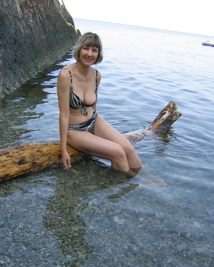 Femme aux cheveux gris platine, en bikini noir et blanc, assise sur un tronc d’arbre dans l’eau claire près d’une falaise rocheuse.