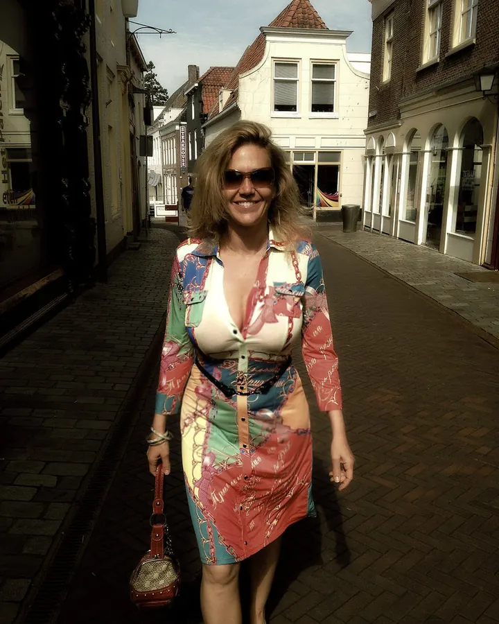 Femme aux cheveux blonds, lunettes de soleil, en robe colorée, marchant dans une rue pavée entre des bâtiments anciens.
