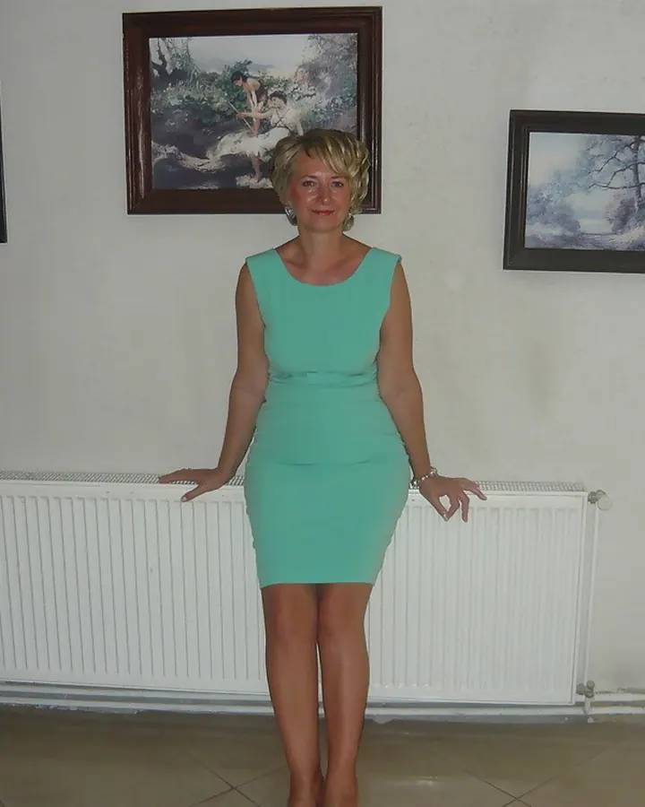 Femme blonde en robe verte, debout devant un radiateur blanc, dans une pièce aux murs clairs.