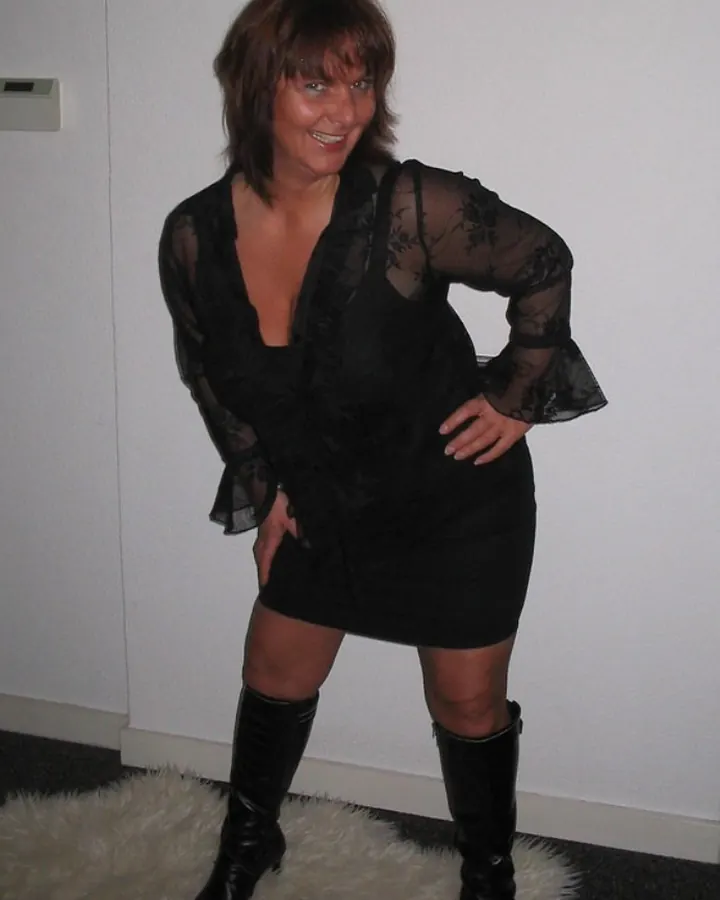 Femme en robe noire transparente, posant sur un tapis blanc, souriante, en bottes noires.