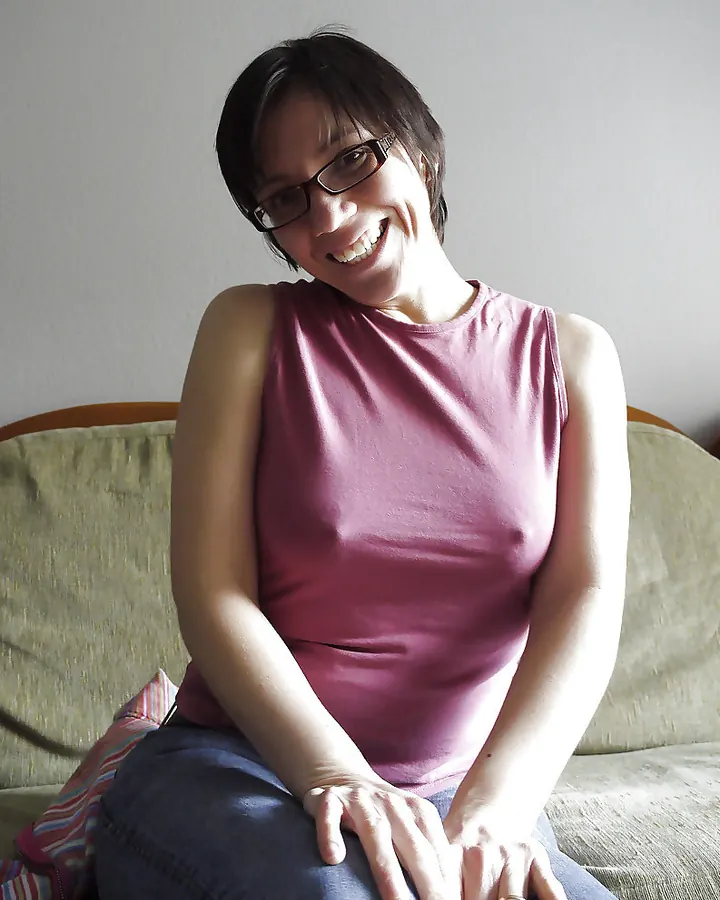 Femme aux cheveux bruns noirs courts, lunettes, souriante, portant un débardeur rose, assise sur un canapé beige.