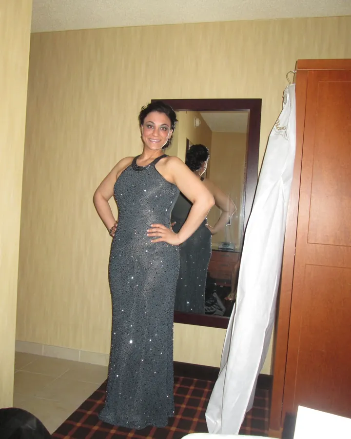 Femme souriante en robe longue argentée pailletée, posant devant un miroir dans une chambre d'hôtel.