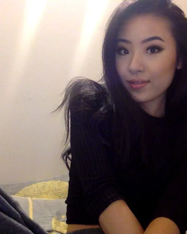 Femme asiatique aux cheveux noirs longs, en pull noir, assise sur un lit avec une lampe à côté.