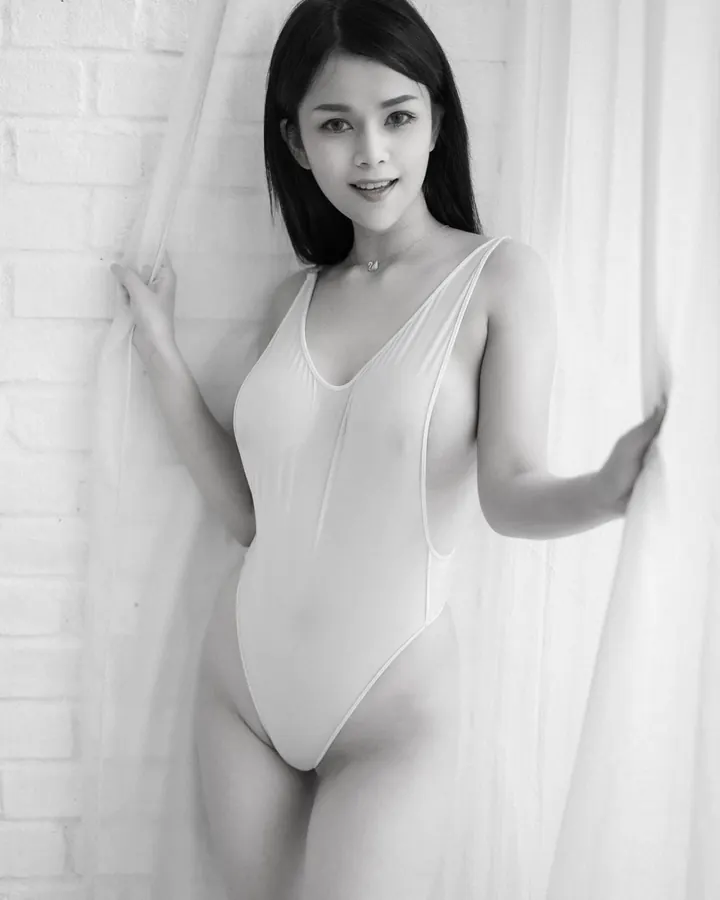 Femme asiatique en maillot blanc, souriante, tenant une fine rideau devant un mur de briques blanches.