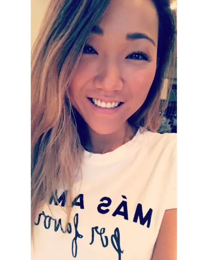 Femme asiatique en lingerie noire, les cheveux foncés, posant sur un lit avec une main dans les cheveux.
