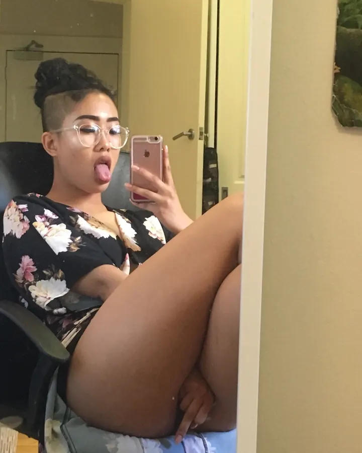 Femme aux cheveux longs portant un haut noir, posant devant une fenêtre avec des volets blancs.
