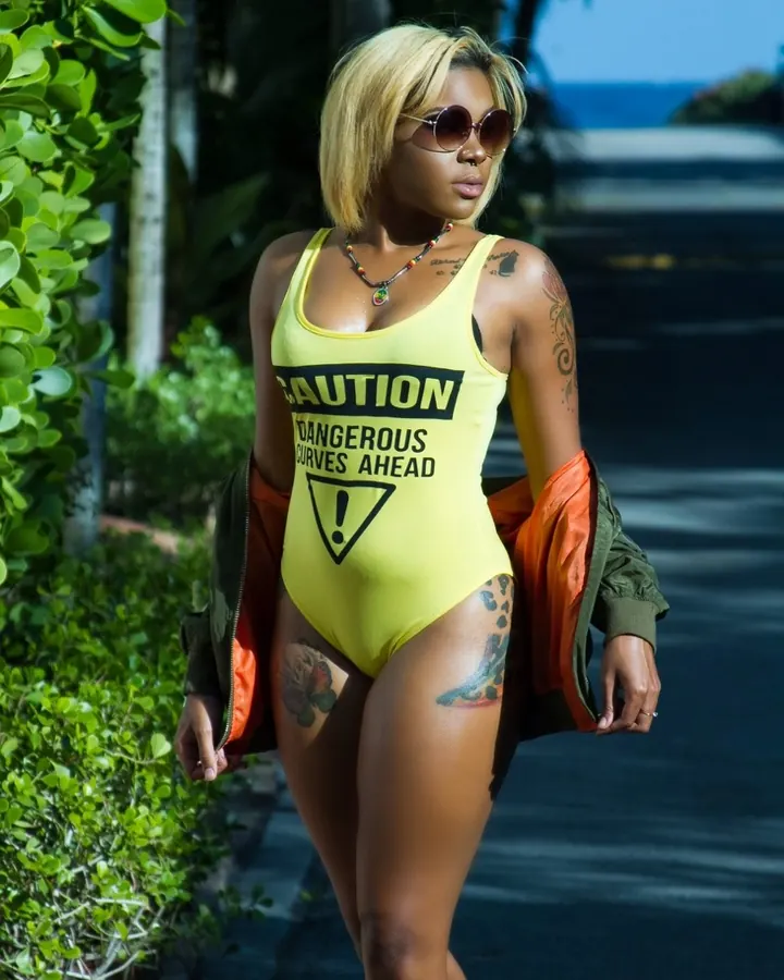 Femme noire aux cheveux blonds, portant un maillot de bain jaune avec l'inscription 'CAUTION DANGEROUS CURVES AHEAD', marchant sur un trottoir.