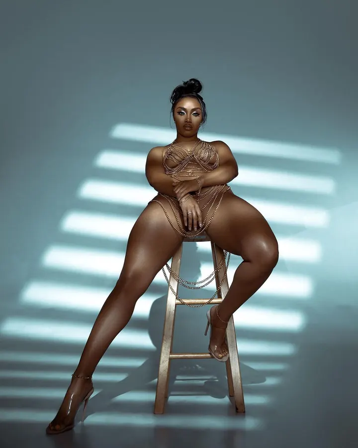 Femme noire assise sur un tabouret, vêtue d'un ensemble doré de chaînes, sous des rayons de lumière horizontaux.