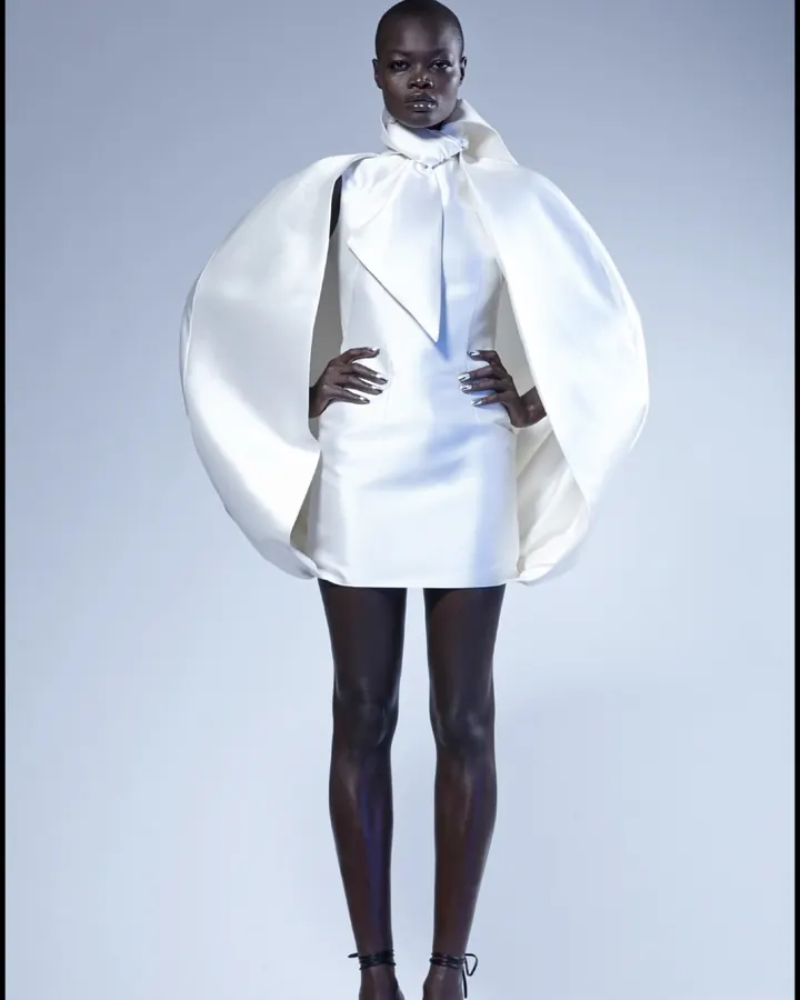 Femme noire aux cheveux ras, portant une robe blanche avec un grand col, posant devant un fond blanc.