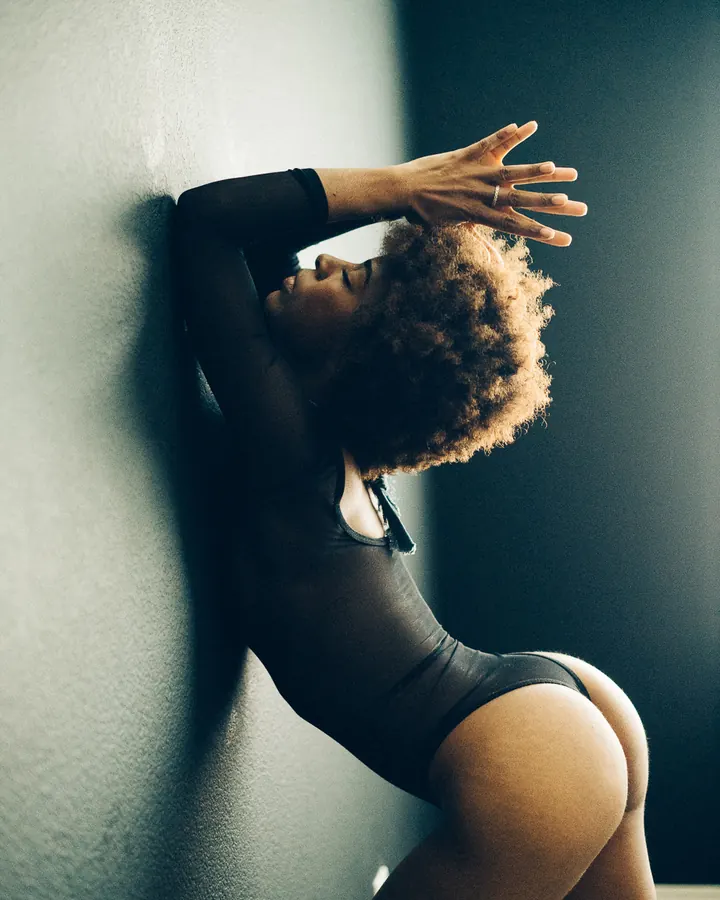Femme noire aux cheveux bouclés portant un body noir, appuyée contre un mur avec les mains derrière la tête.
