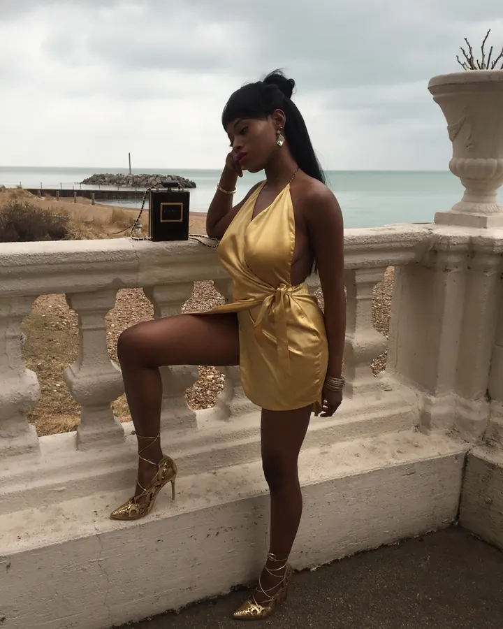 Femme noire aux longs cheveux tressés, portant une robe rose transparente, assise sur une chaise blanche.