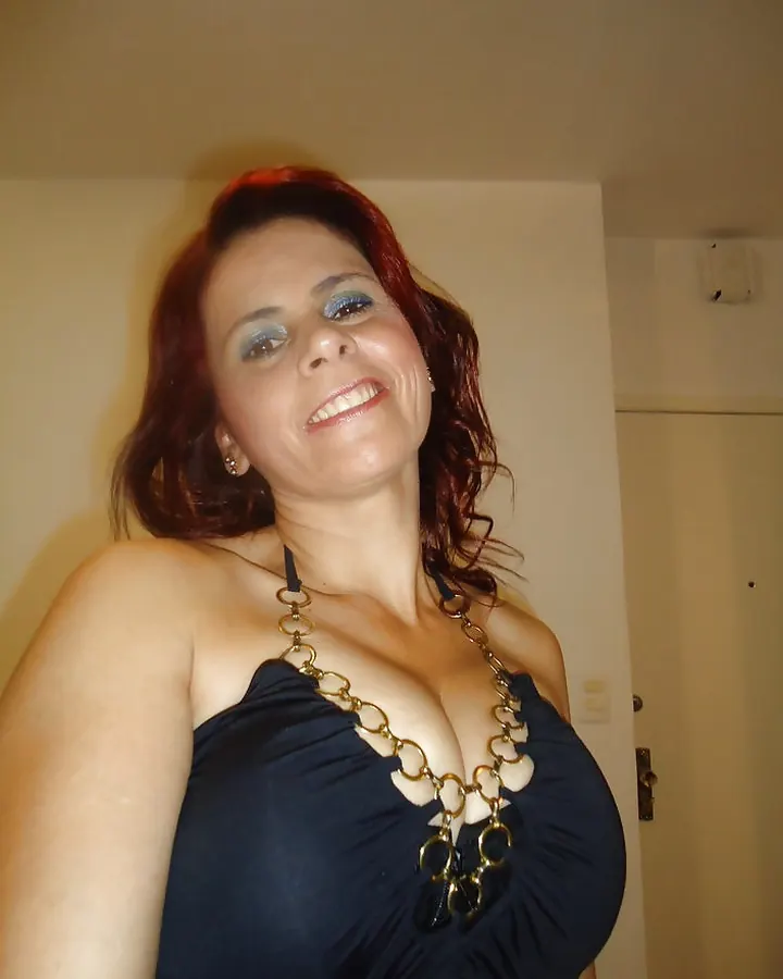 Femme aux cheveux roux, souriante, portant un haut noir à chaînes dorées, décolleté profond.