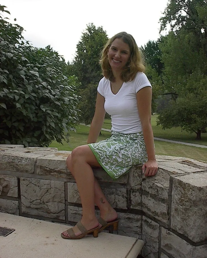 Femme souriante, assise sur un mur de pierre, portant un t-shirt blanc et une jupe verte à motifs, sandales à talons.