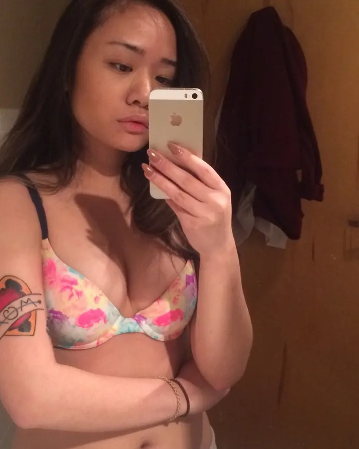 Jeune femme asiatique en soutien-gorge fleuri prenant un selfie dans une salle de bain, tatouage visible sur le bras.