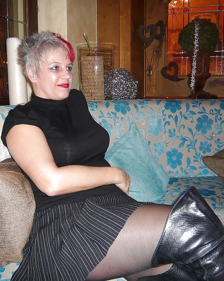 Femme grise en robe noire assise sur un canapé, portant des collants et des bottes en cuir.