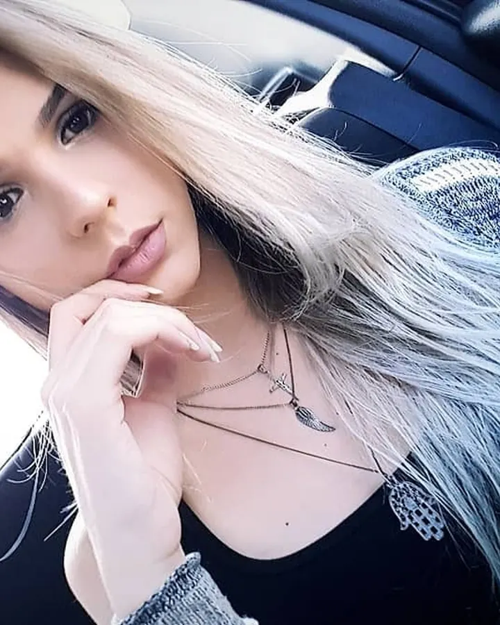 Femme blonde en pull gris assise dans une voiture, regardant l'appareil photo, cheveux longs et ongles manucurés.