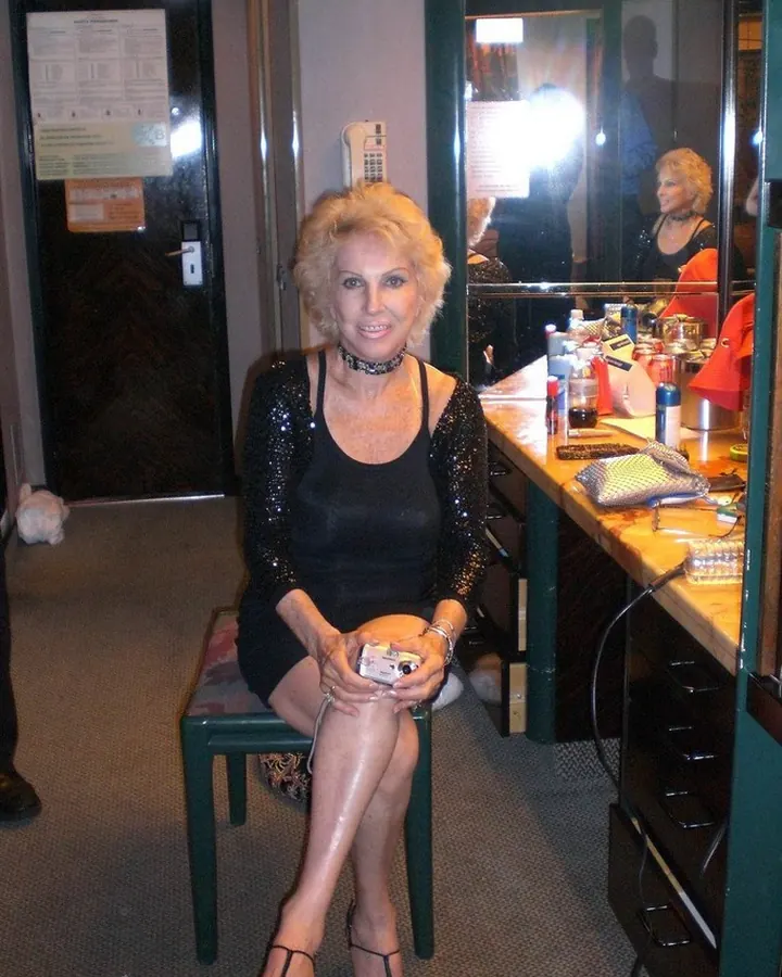 Femme blonde aux yeux marrons, souriante, assise sur une chaise verte en tenue noire scintillante