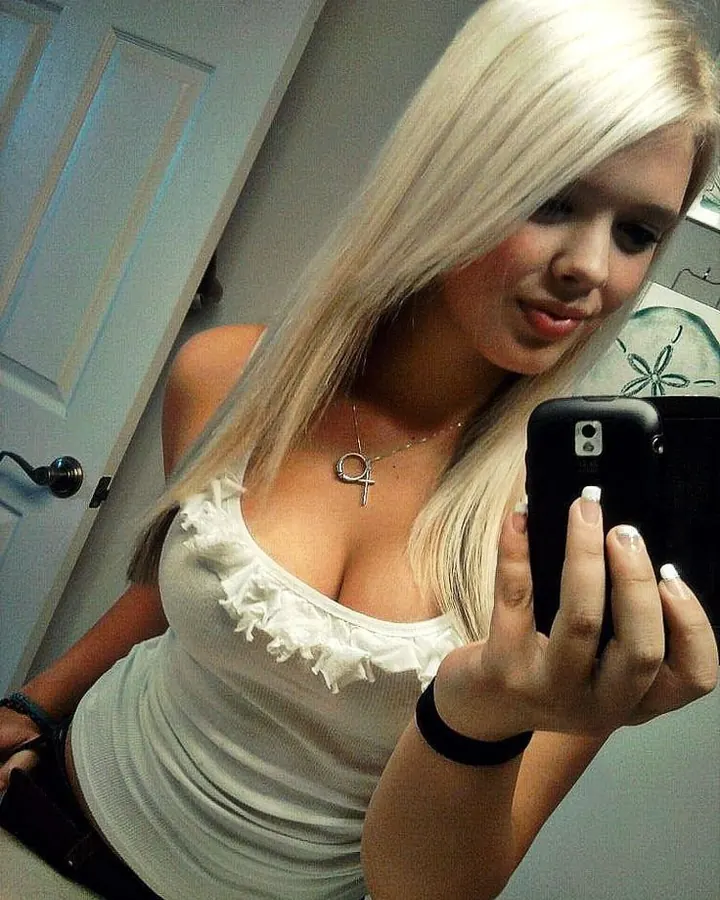 Femme aux cheveux platine en tenue blanche, tenant un smartphone, dans une chambre avec un mur en bois