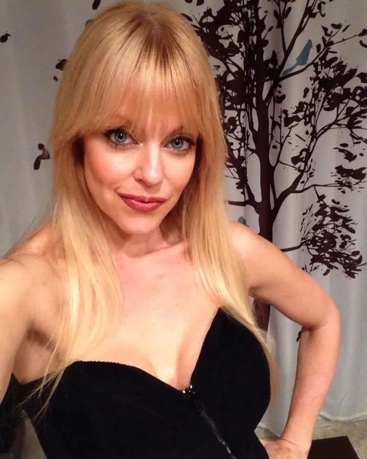 Femme aux cheveux blonds en tenue blanche, assise sur un canapé blanc dans une pièce sombre