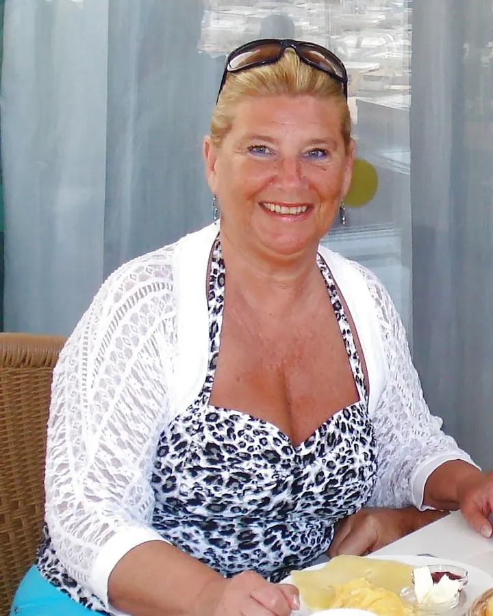 Femme blonde souriante portant un haut à motif léopard et une veste blanche, assise à une table avec une assiette de nourriture