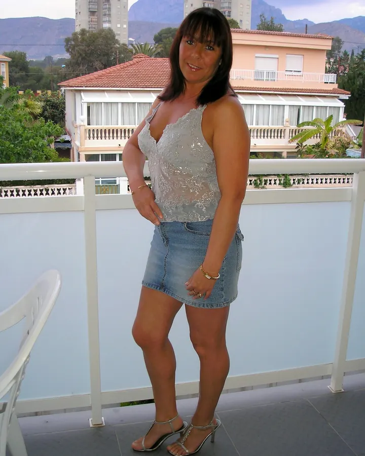Femme aux cheveux bruns portant une robe transparente et une jupe en jean, debout sur un balcon avec vue sur des bâtiments et des montagnes