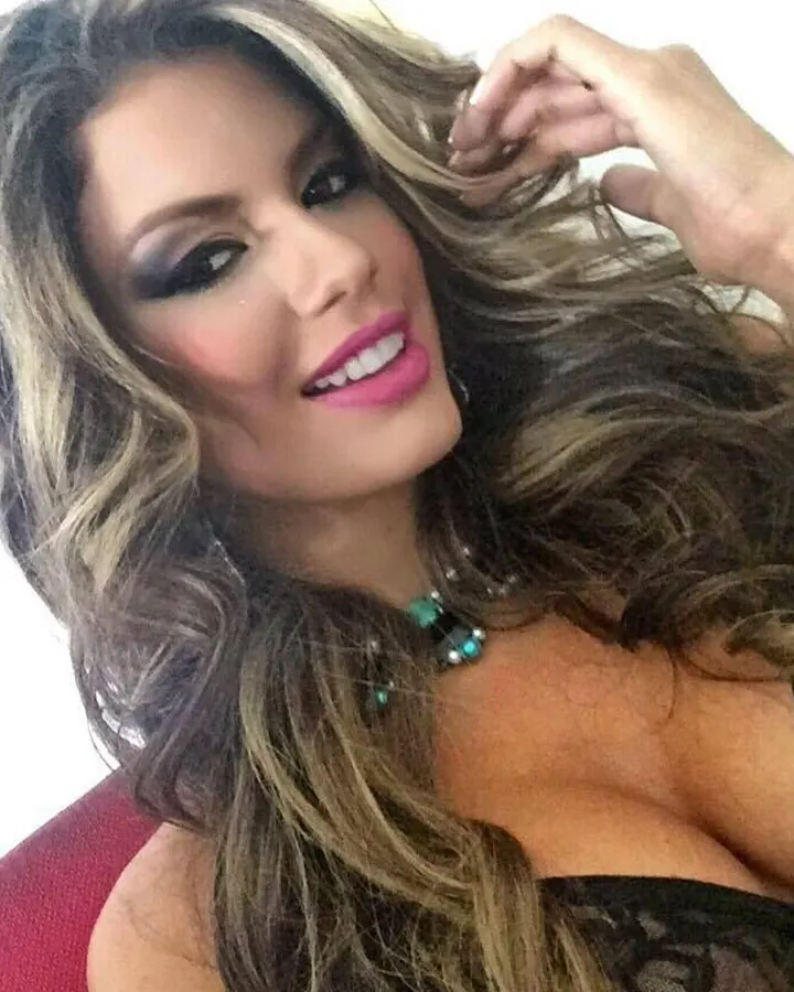 Femme aux cheveux blonds ondulés, maquillage prononcé, souriant à l'appareil photo, avec un collier turquoise, sur fond rouge.