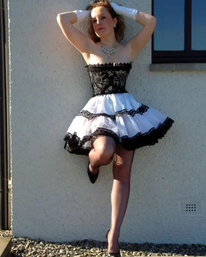 Femme aux cheveux châtains, vêtue d'un blazer marron et d'une jupe noire en cuir, debout sur un sol brillant, avec des collants transparents et des talons noirs.