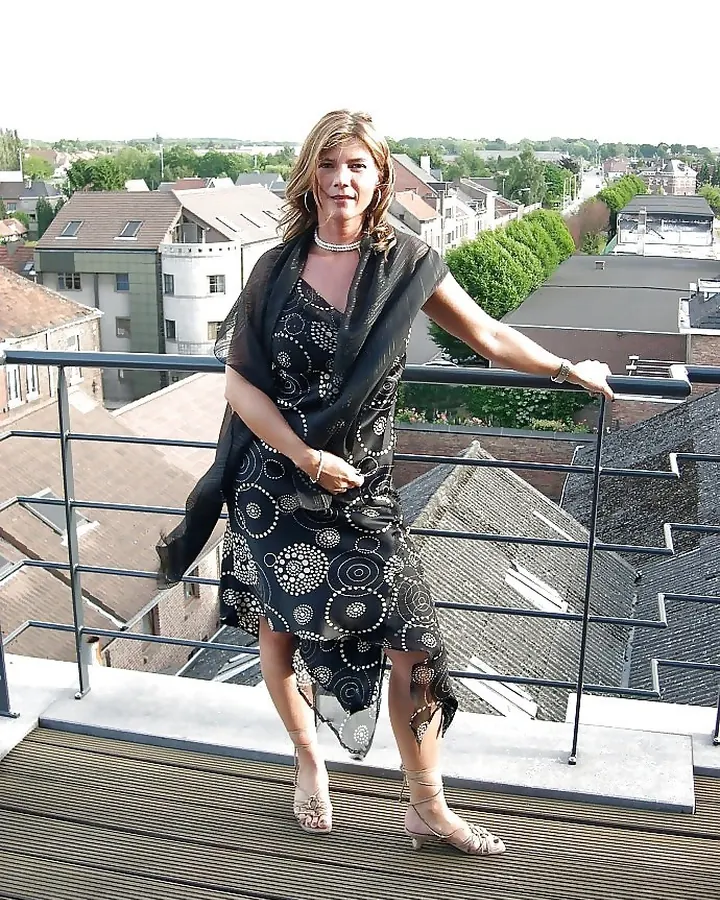 Femme blonde, vêtue d'un haut noir à lacets, posant de profil sur fond blanc, avec un sourire discret.