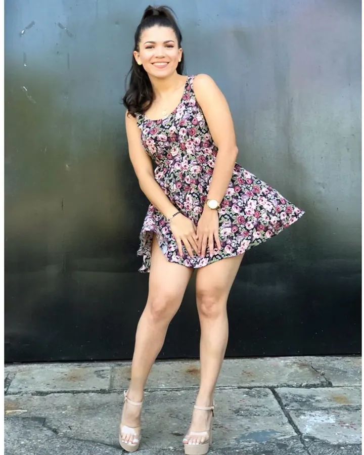 Femme souriante en robe fleurie courte, debout contre un mur sombre sur un trottoir pavé, les mains posées sur ses cuisses.