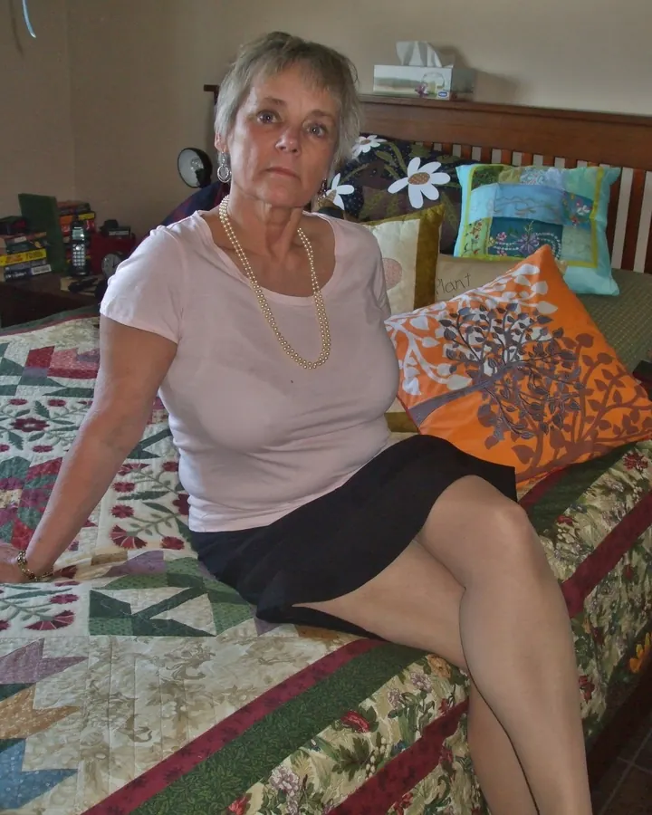 Femme aux cheveux gris courts, vêtue d'un t-shirt rose et d'une jupe noire, assise sur un lit coloré avec coussins décoratifs.