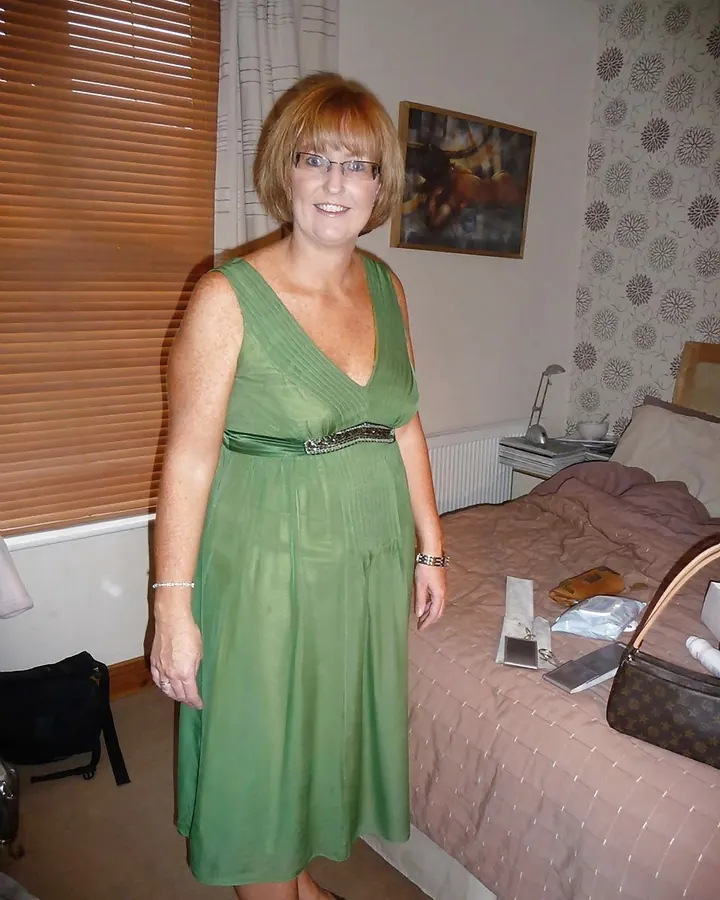 Femme aux cheveux châtains courts, lunettes, robe verte avec ceinture, souriante dans une chambre avec lit et rideaux.