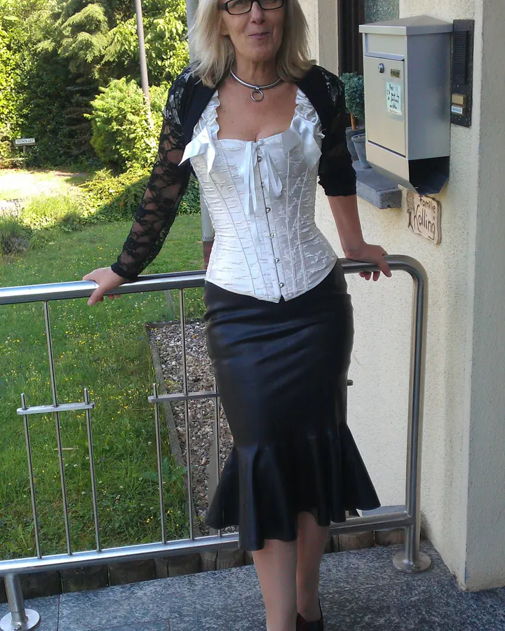 Femme aux cheveux blancs courts, souriante, vêtue d’un haut blanc à lacets, cardigan noir transparent, jupe noire en cuir, lunettes, sur un balcon.