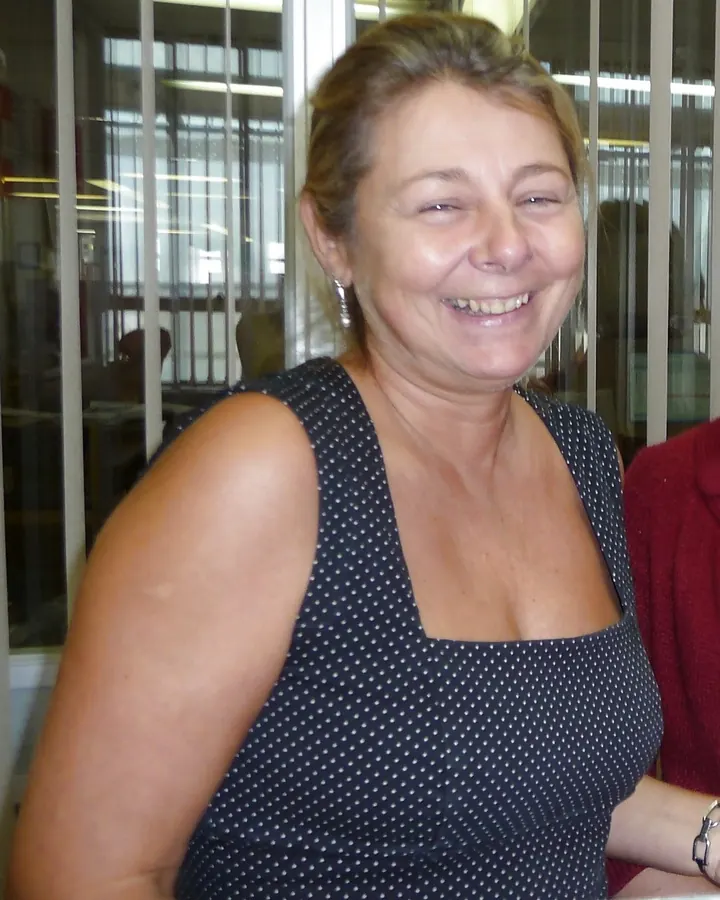 Femme aux cheveux blonds courts, souriante, vêtue d’un haut noir en dentelle transparent, gants longs noirs, mains sur les hanches.