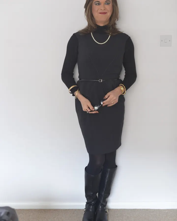 Femme aux cheveux blonds mi-longs, souriante, vêtue d’une robe noire ajustée avec col roulé, ceinture fine, collants noirs et bottes hautes.