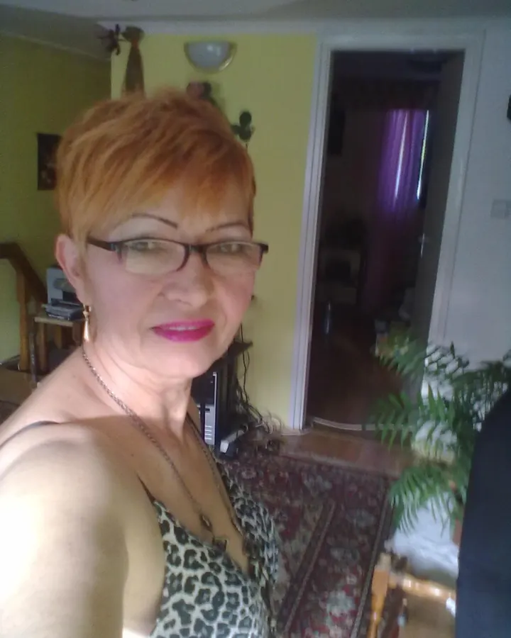 Femme aux cheveux roux courts, portant des lunettes et un haut léopard, souriant, dans un salon aux murs jaunes.