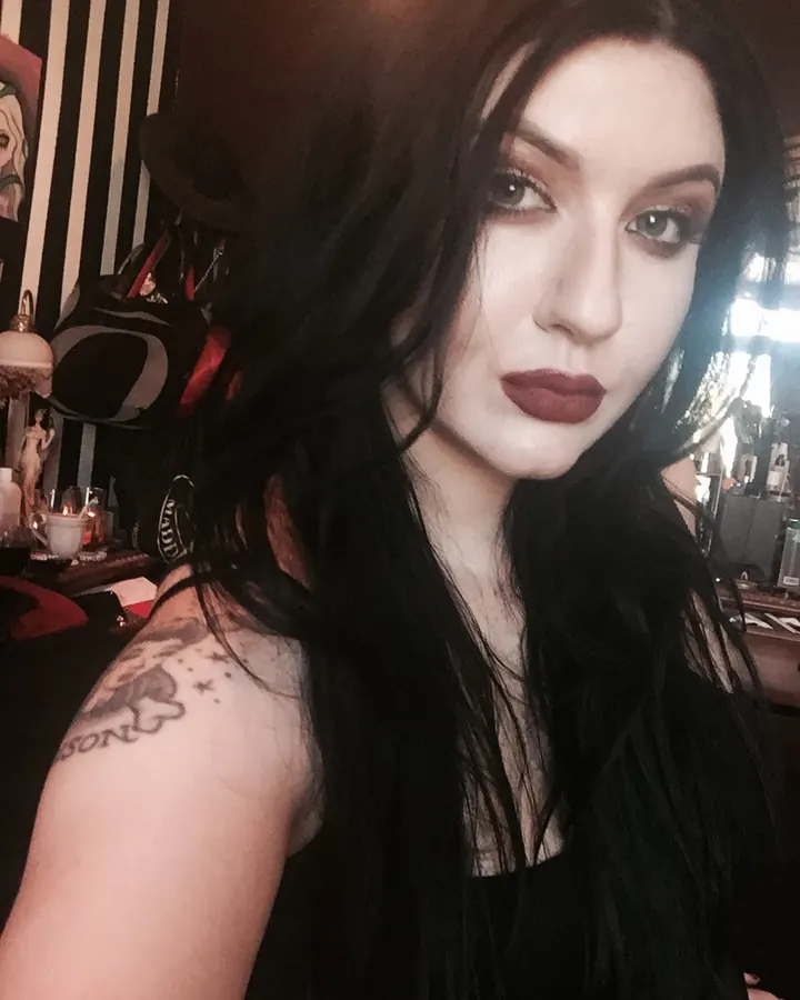 Femme aux cheveux bruns, vêtue d'une tenue noire, posant devant un mur, mains sur les hanches, regard direct.